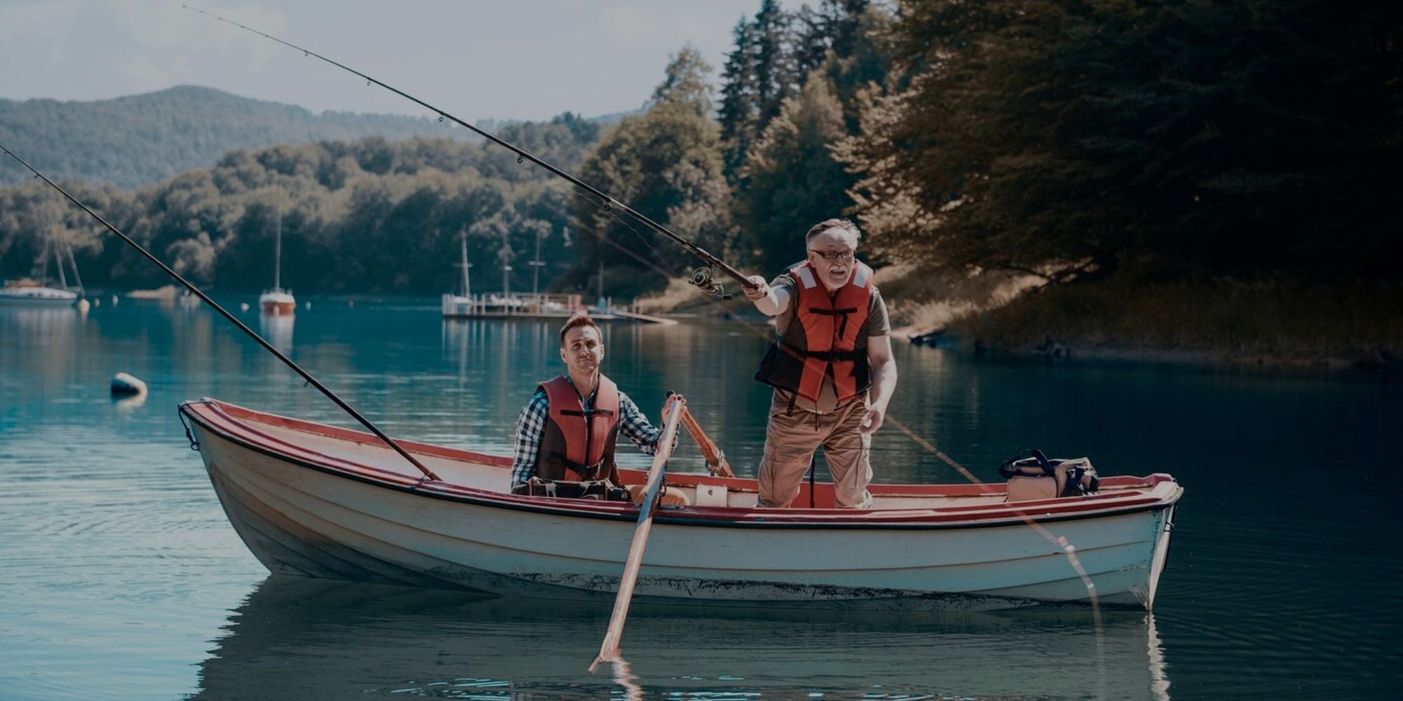 Two people in a small white and red boat on a calm lake, each wearing an orange life vest and holding a fishing rod, with a forested shoreline in the background.