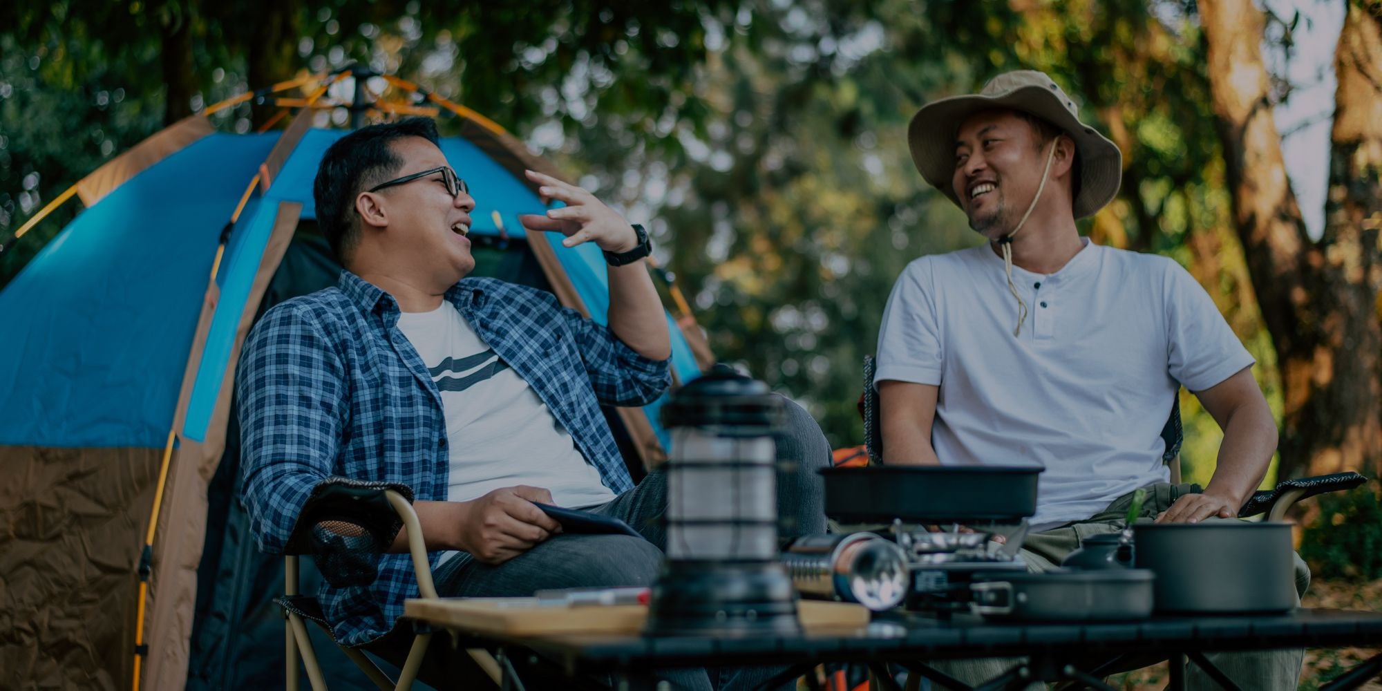 Two men camping outdoors next to a blue and brown tent, with one wearing a blue plaid shirt and the other wearing a white t-shirt and a wide-brimmed hat. A camping stove, lantern, and pots are on a table in the foreground.